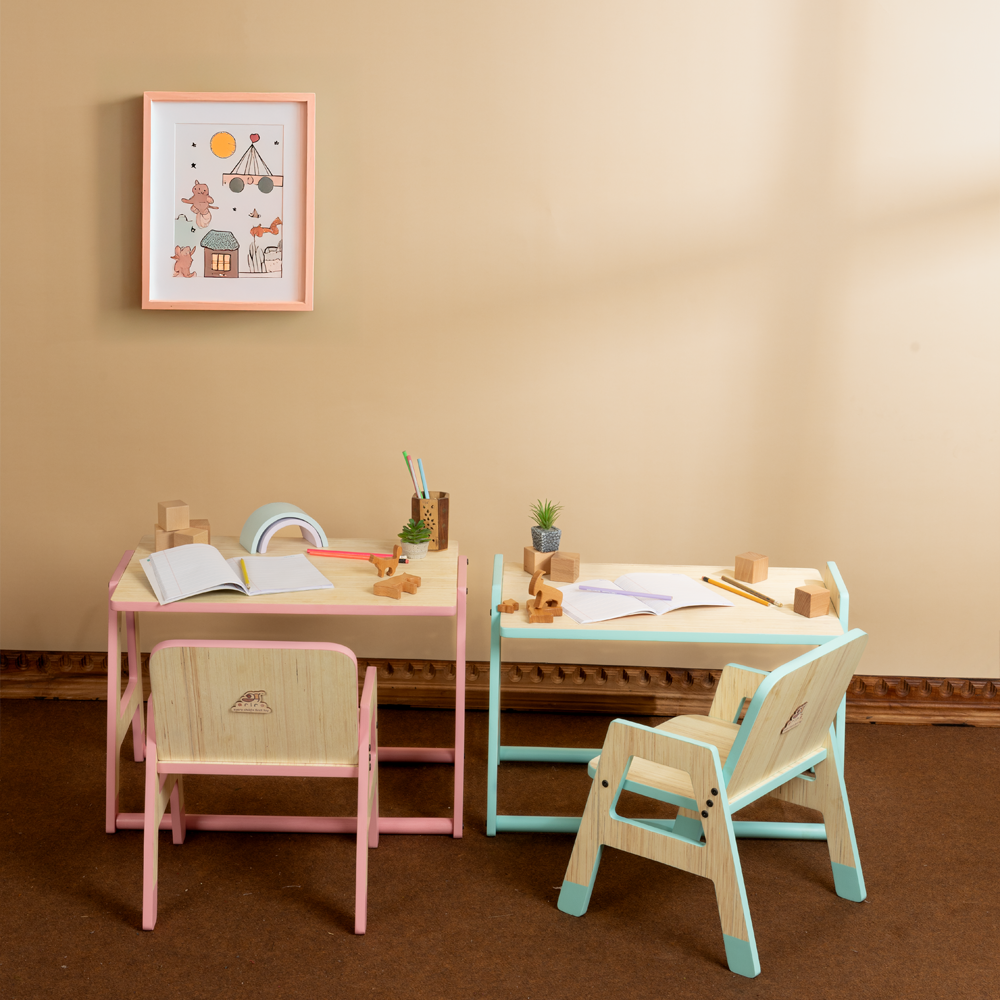 Two wooden toddler table and chair sets in pink and mint-green color options, styled side by side with books, plants, and wooden toys, under a framed illustration on the wall.