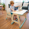 Young boy sitting at a mint-green wooden toddler table and chair, turning the pages of a picture book.