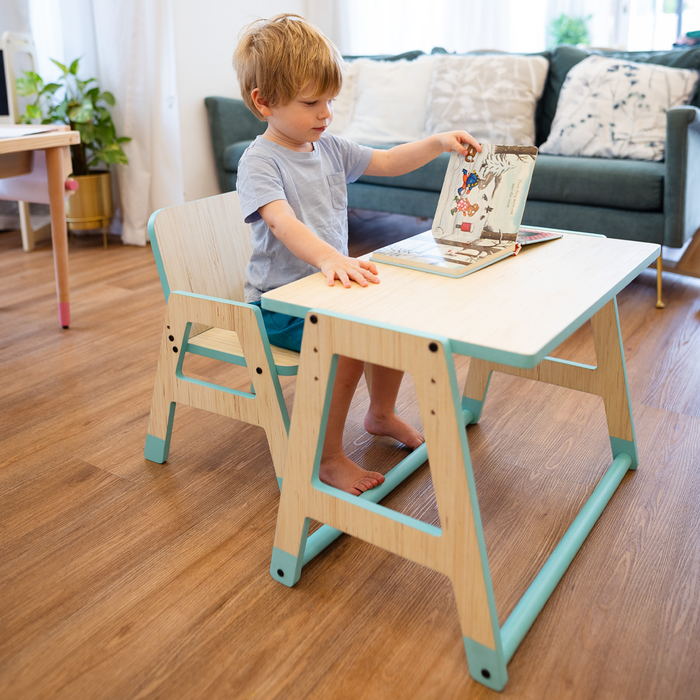 Young boy sitting at a mint-green wooden toddler table and chair, turning the pages of a picture book.