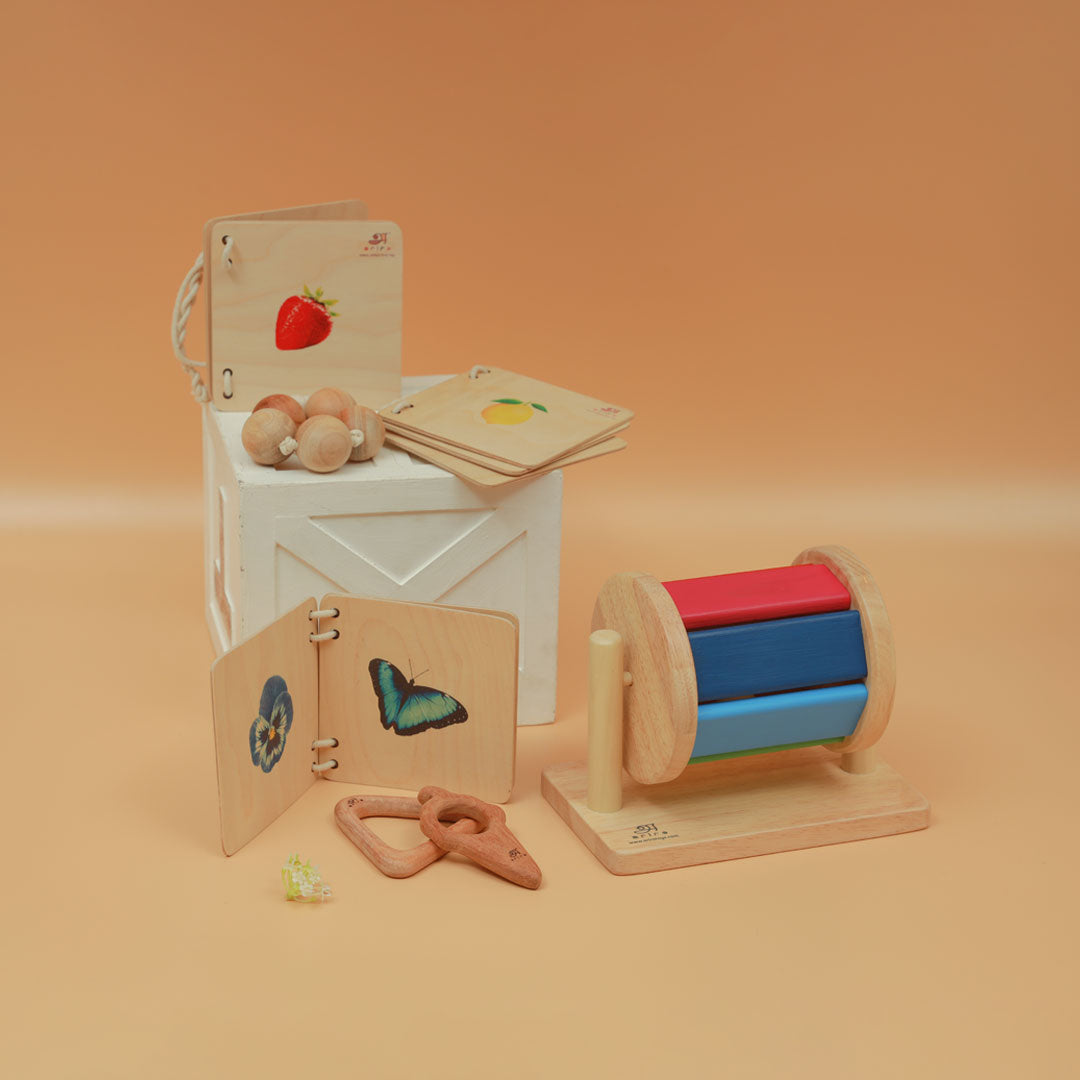 Montessori Kit i has nteractive books with colorful illustrations, grasping beads, a wooden teether, and a spinning drum, all designed to encourage sensory and motor skill development.