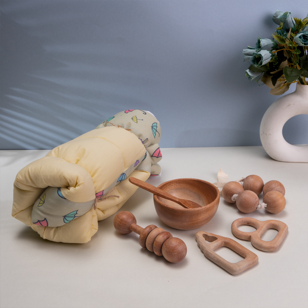 new born gift set include a rolled-up yellow blanket with a colorful umbrella pattern, a small wooden bowl and spoon, a wooden dumbbell rattle, a set of grasping beads, and a wooden teething ring.