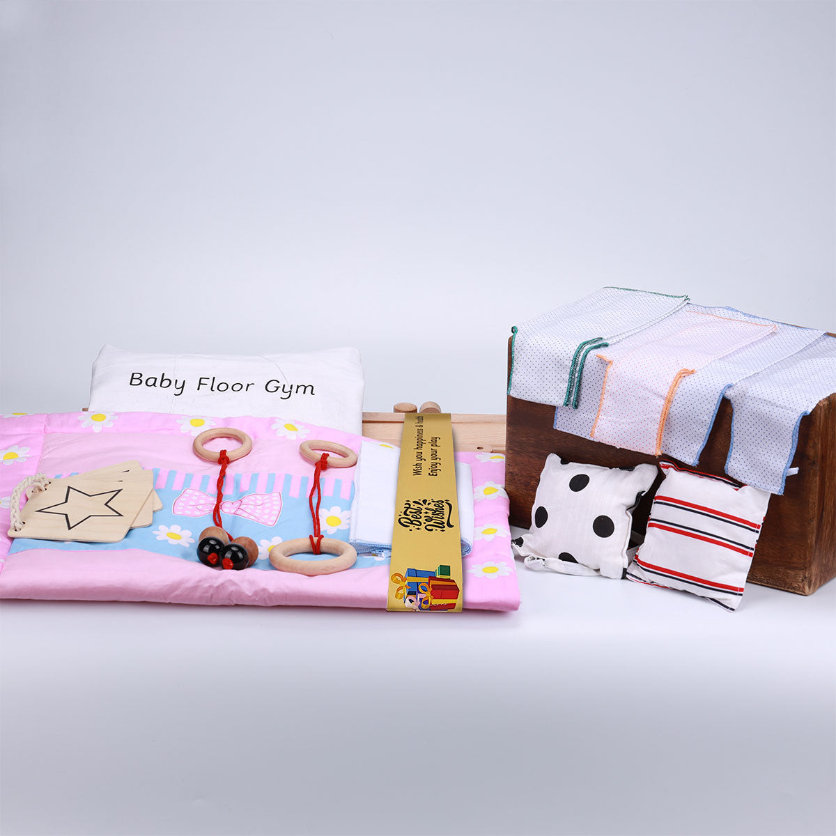 A baby floor gym set consist a pink play mat, a pillow, wooden hanging toys, and a selection of cloths and cushions, perfect for a child's playtime.