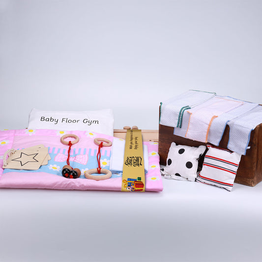 A baby floor gym set consist a pink play mat, a pillow, wooden hanging toys, and a selection of cloths and cushions, perfect for a child's playtime.